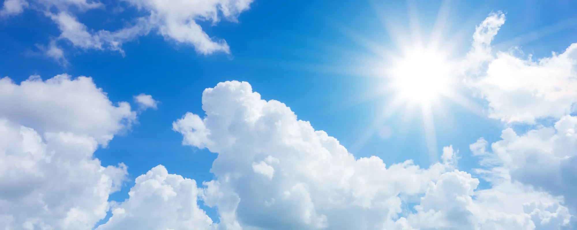 Ein strahlend blauer Himmel mit leuchtend weißer Sonne, die ihre wärmenden Strahlen ausbreitet. Fluffige, weiße Wolken schweben sanft über den Himmel, und das Licht erzeugt eine helle, klare Atmosphäre. Die Szene vermittelt ein Gefühl von Weite, Freiheit und Leichtigkeit.