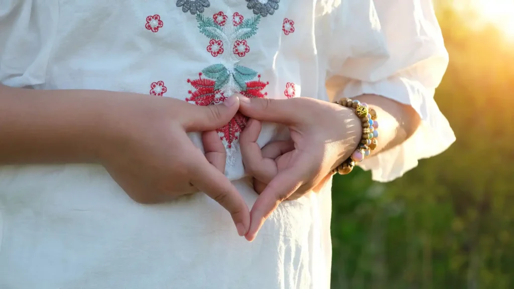 Eine Person in einer weißen, bestickten Bluse formt mit den Händen ein Mudra, eine symbolische Handgeste. Die Finger sind sanft aneinandergelegt und bilden eine Herzform. Am rechten Handgelenk sind bunte Perlenarmbänder zu sehen. Im Hintergrund scheint die Sonne warm und golden durch das Grün der Natur.