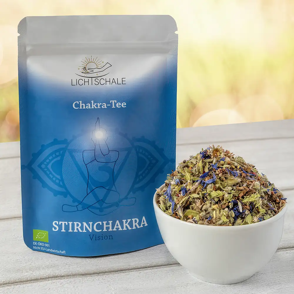 Blau-violette Teeverpackung mit der Aufschrift ‚Chakra-Tee Stirnchakra – Vision‘ von Lichschale. Daneben eine weiße Schale mit losem, blumigem Kräutertee, auf einem hellen Holztisch vor unscharfem Naturhintergrund