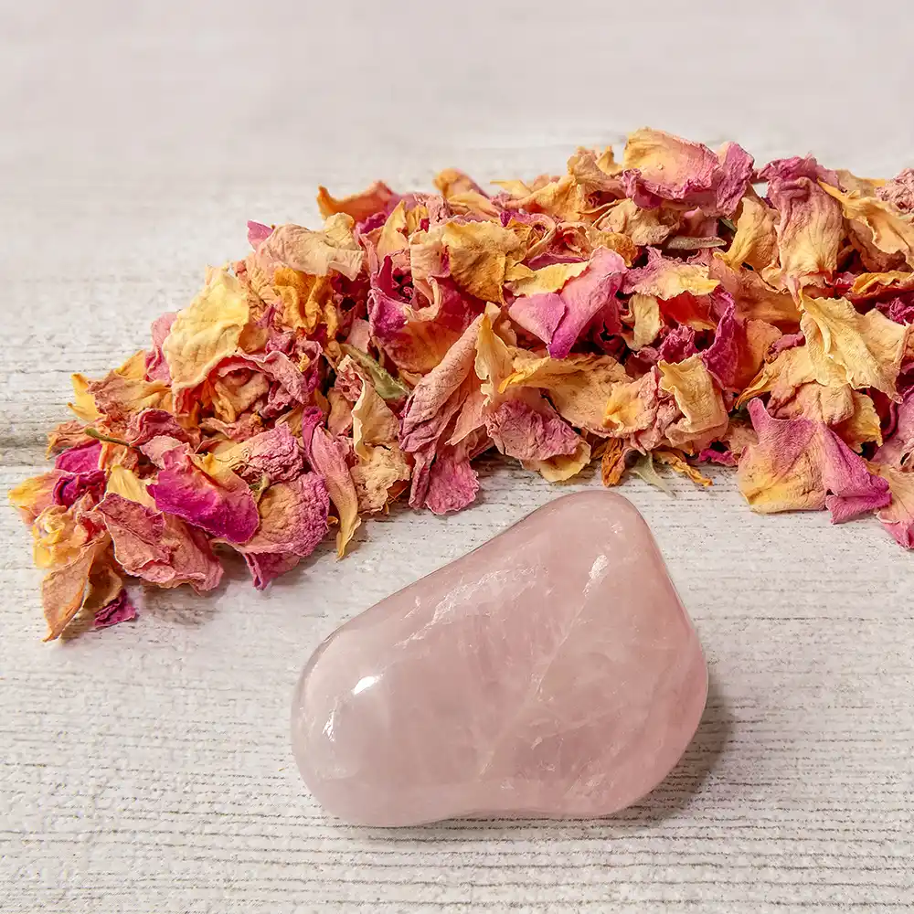 Zartrosafarbener, polierter Rosenquarz-Stein vor einem Haufen getrockneter, rosa-gelber Rosenblüten auf hellem Holzuntergrund