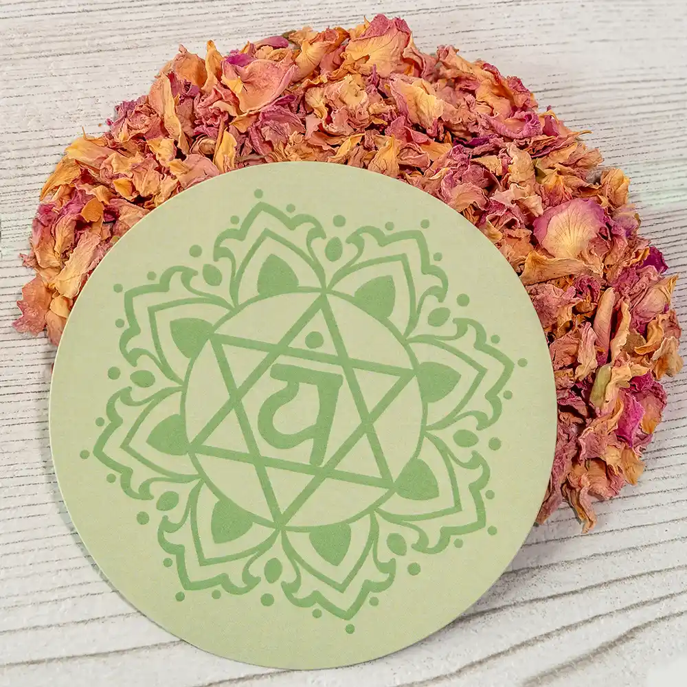 Grüner, runder Untersetzer mit dem Symbol des Herzchakras (Anahata), liegend auf hellem Holz, umgeben von rosa und gelben getrockneten Blütenblättern