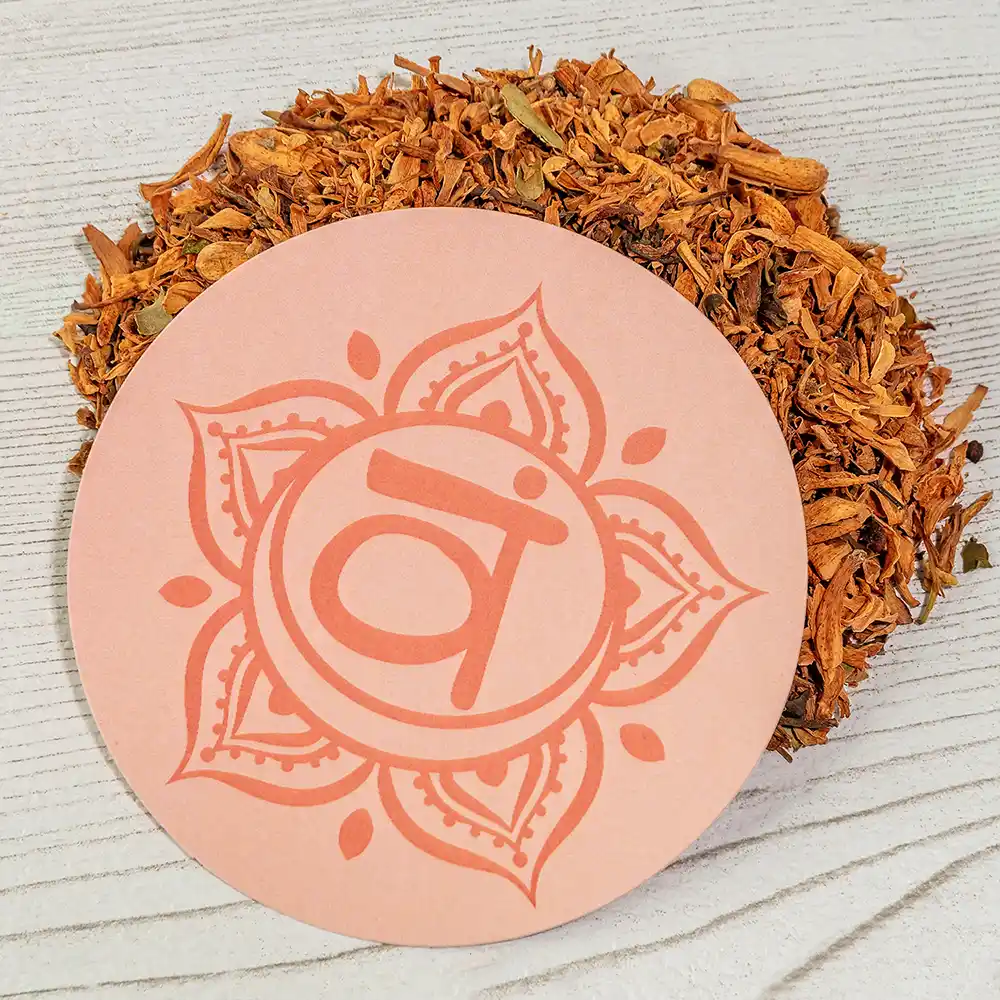 Runder, orangefarbener Untersetzer mit dem Symbol des Sakralchakras (Svadhisthana), platziert auf hellem Holz und umgeben von getrockneten, orange-braunen Kräutern