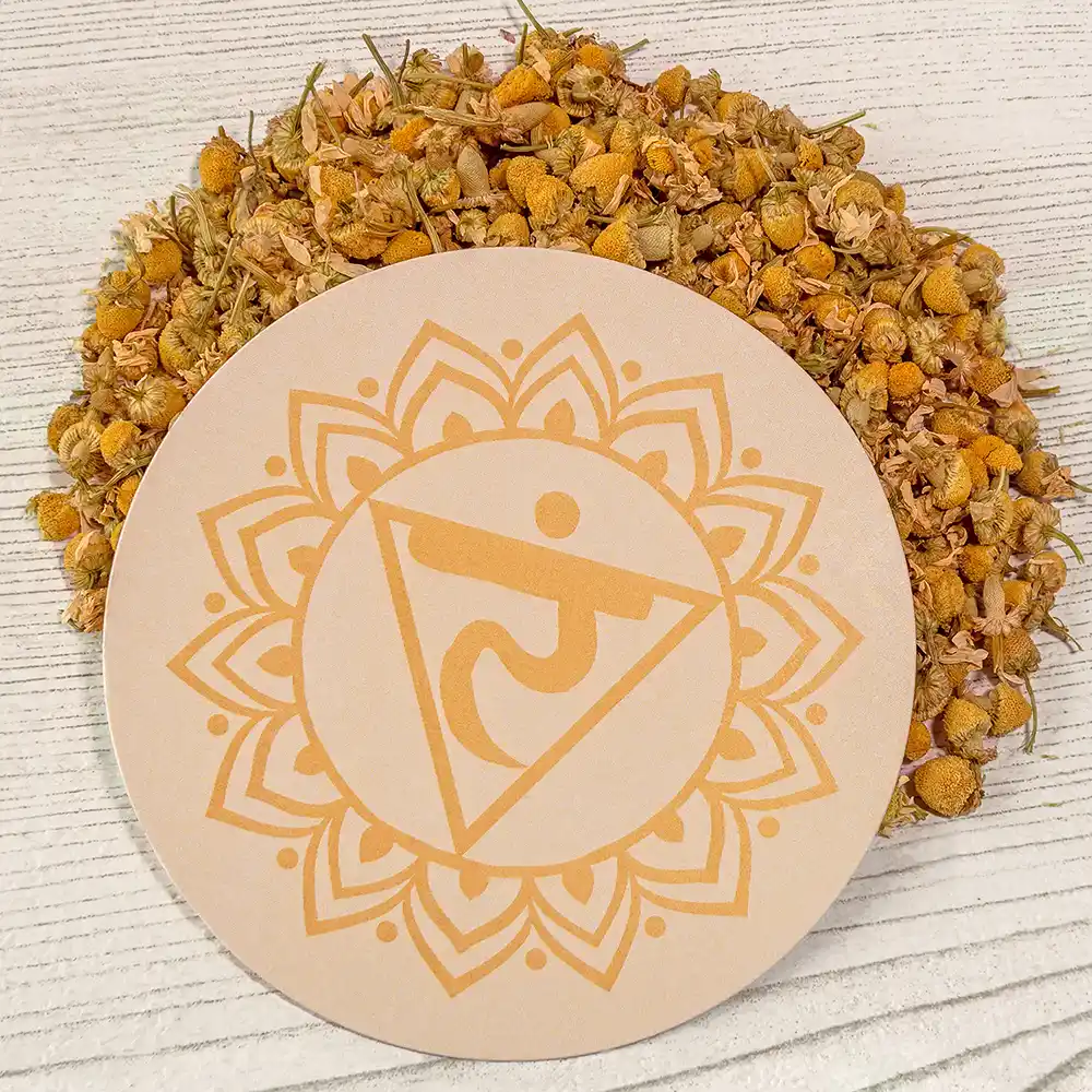 Runder, gelber Untersetzer mit dem Symbol des Solarplexus-Chakras (Manipura), liegend auf hellem Holz, umgeben von getrockneten Kamillenblüten