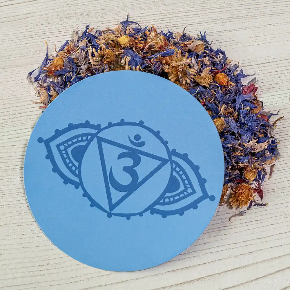Ein runder, blauer Untersetzer mit dem Symbol des Stirnchakras (Ajna) liegt auf hellem Holz, umgeben von getrockneten, blau-violetten Blüten