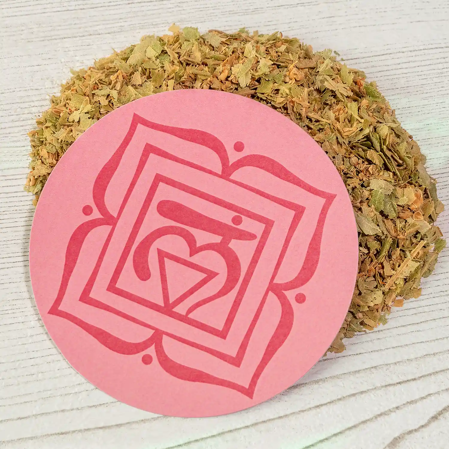 Ein runder, rosaroter Untersetzer mit dem Symbol des Wurzelchakras (Muladhara) liegt auf hellem Holz, umgeben von getrockneten, grünlich-braunen Kräutern