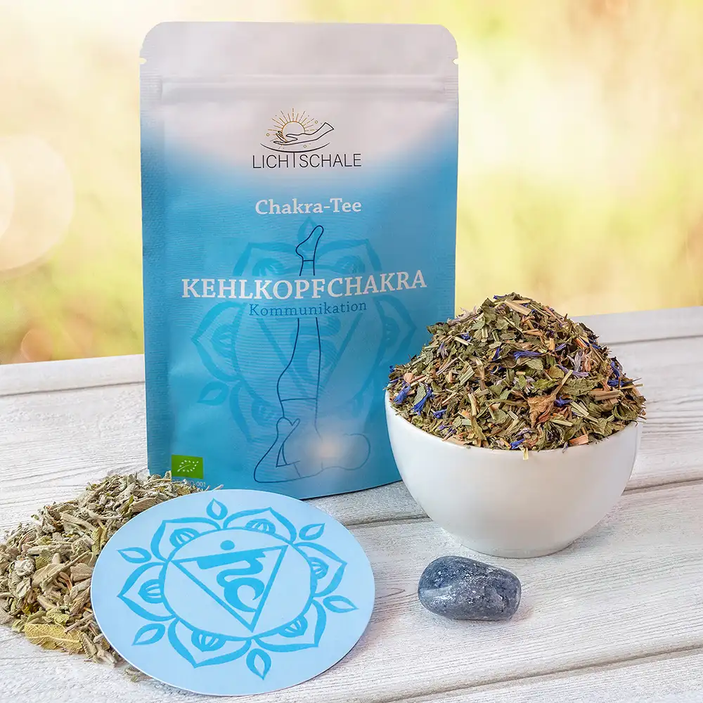 Chakra-Tee-Set für das Kehlkopfchakra mit hellblauer Verpackung, Kräutermischung in weißer Schale, losem Tee, runder Unterlage mit Vishuddha-Symbol und einem blaugrauen Heilstein auf Holzuntergrund