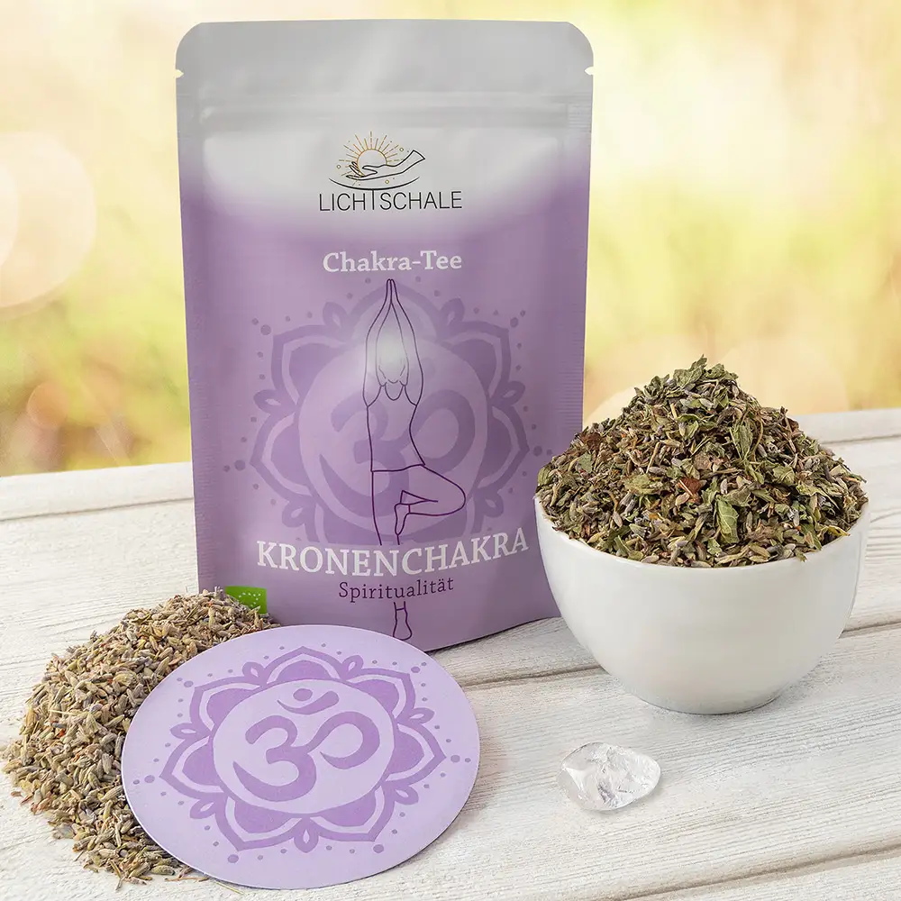 Chakra-Tee-Set für das Kronenchakra mit violetter Verpackung, losen Teekräutern in einer weißen Schale, Lavendelblüten, einer Unterlage mit Om-Symbol und einem kleinen Kristall auf hellem Holztisch