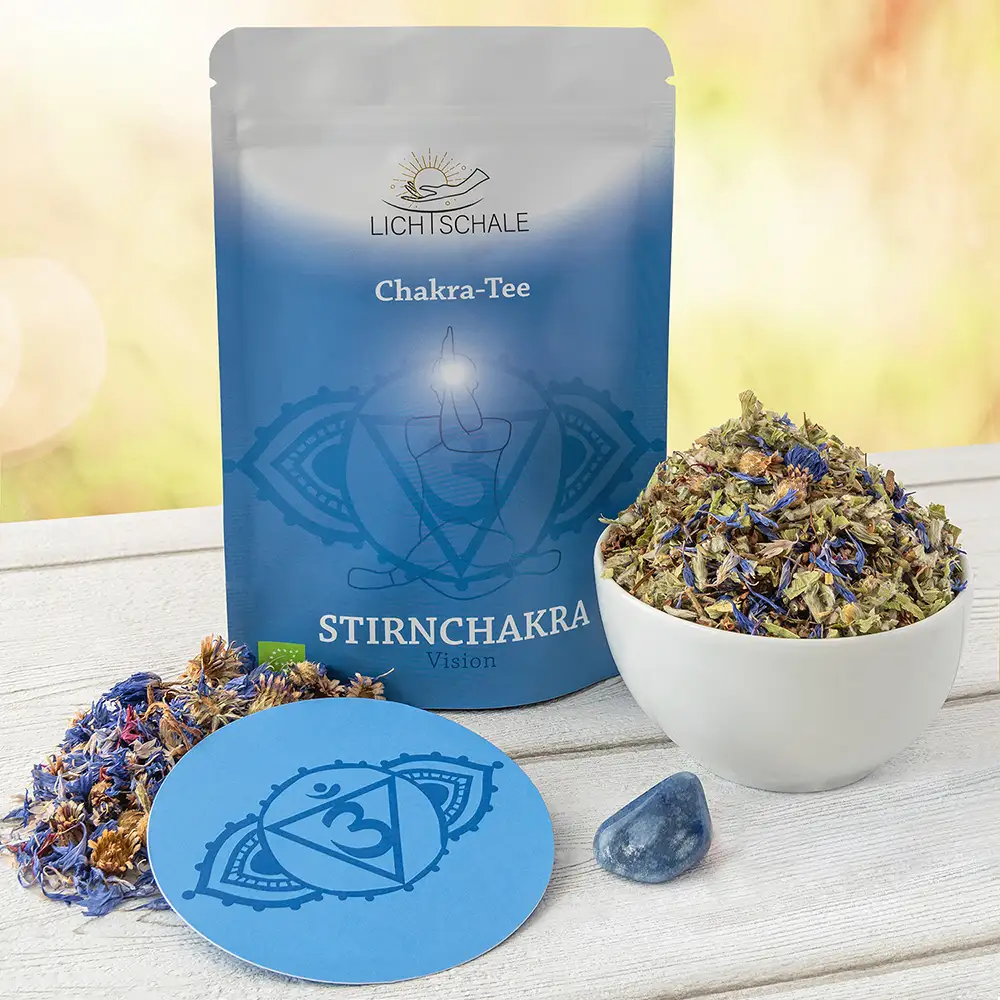 Chakra-Tee-Set für das Stirnchakra mit blauer Verpackung, losem Kräutertee in weißer Schale, blauen Blüten, einem Untersetzer mit Ajna-Symbol und einem dunkelblauen Edelstein auf hellem Holztisch